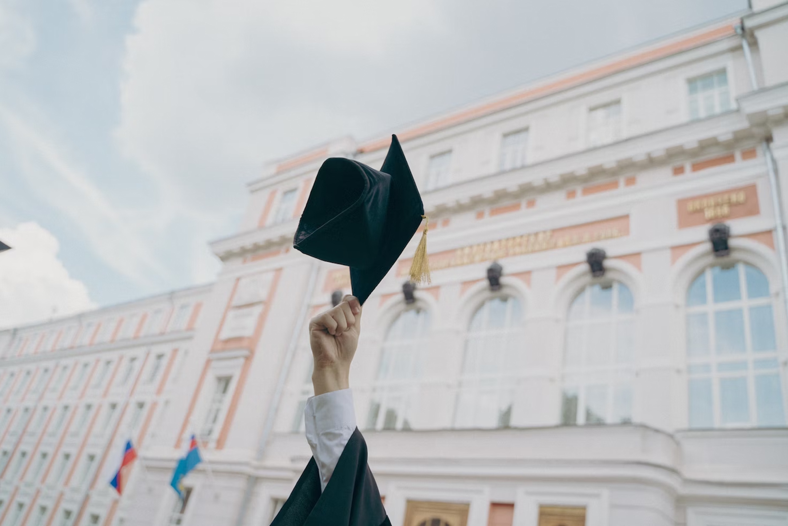 A recently graduated student holds up their graduation cap in front of a school building.