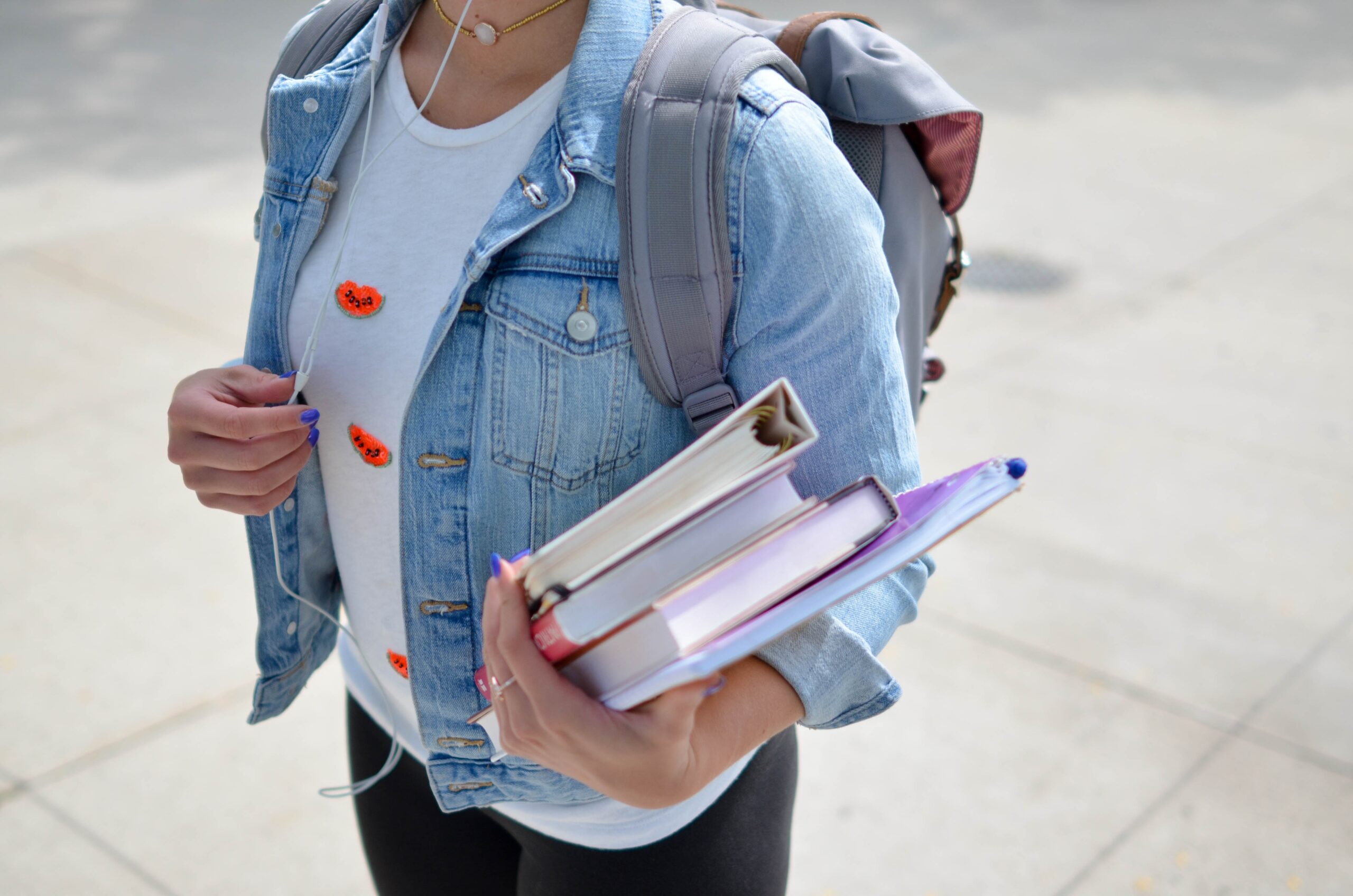 A woman wearing a white shirt and a denim jacket has a backpack on her pack and holds three notebooks in her left arm.