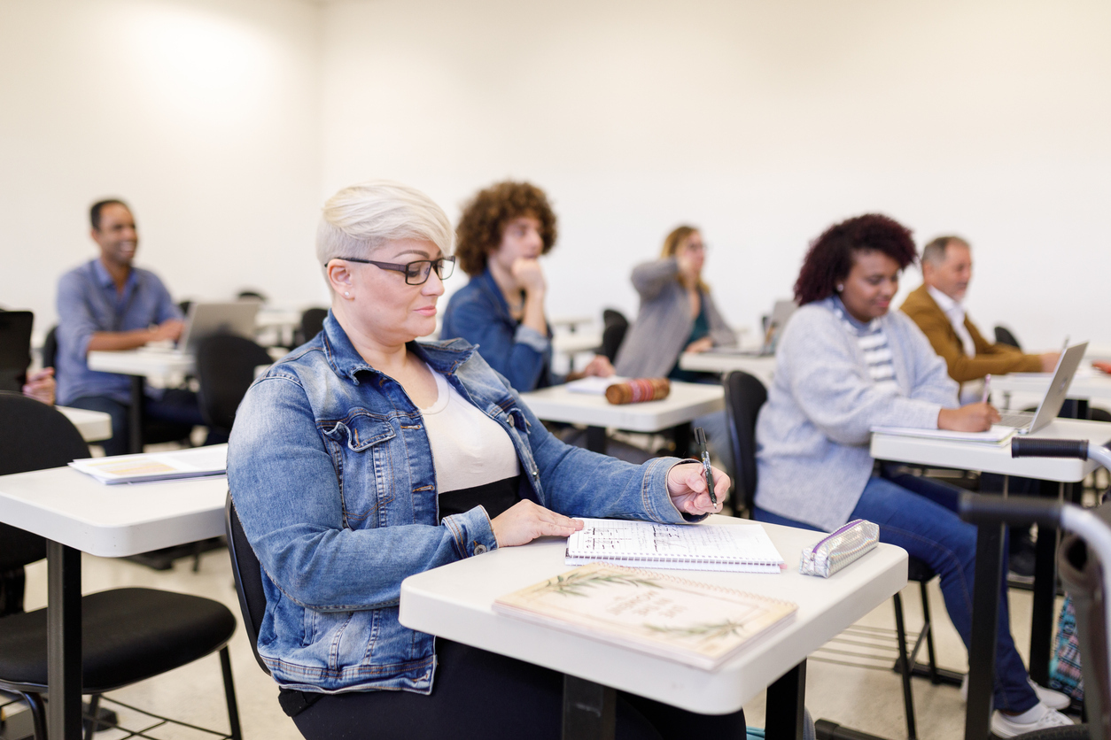 An adult woman with short white hair sits at a desk in a classroom surrounded by other adult learners.